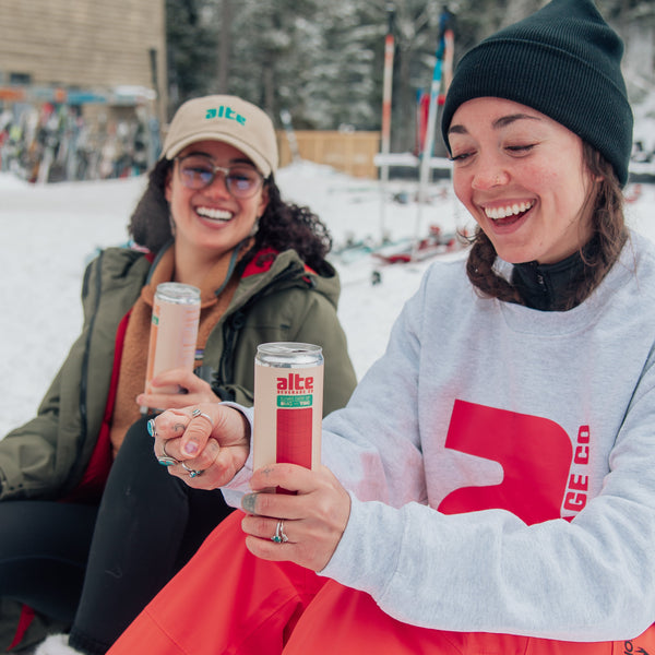 Two people sitting in the snow, holding AlteBev drinks with visible branding.
