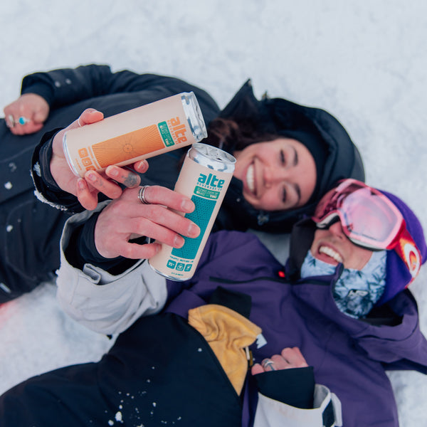 Two people lying in the snow, one holding a can of beer, with a snowy landscape in the background.